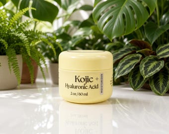 May include: A pale yellow cylindrical jar of moisturizer with a screw-top lid. The label features black text reading Kojic + Hyaluronic Acid, 2 oz / 60 ml, and a vertical strip labeled MOISTURIZER. The jar sits on a reflective white surface.