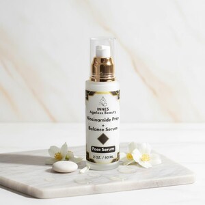 Puede incluir: Un frasco blanco de s&eacute;rum facial con detalles dorados y una tapa transparente. La etiqueta dice "INNES Ageless Beauty Niacinamide Prep + Balance Serum Face Serum 2 OZ. / 60 ML." El frasco est&aacute; sobre una superficie de m&aacute;rmol, con flores blancas y una peque&ntilde;a piedra.