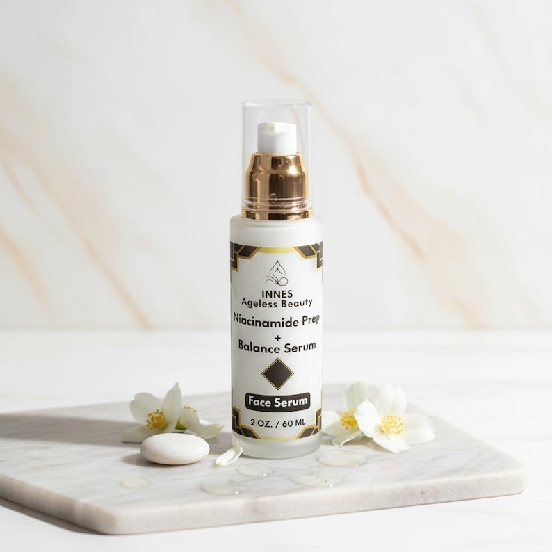 Puede incluir: Un frasco blanco de s&eacute;rum facial con detalles dorados y una tapa transparente. La etiqueta dice "INNES Ageless Beauty Niacinamide Prep + Balance Serum Face Serum 2 OZ. / 60 ML." El frasco est&aacute; sobre una superficie de m&aacute;rmol, con flores blancas y una peque&ntilde;a piedra.