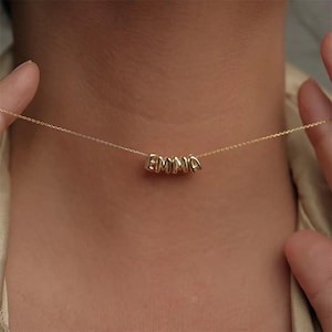 Könnte beinhalten: Eine zarte goldene Halskette mit dem Namen "EMMA", der in einzelnen, abgerundeten Buchstabenperlen geschrieben ist. Die Halskette wird um den Hals getragen, wobei die Buchstaben zentriert sind. Kette und Buchstaben scheinen aus Gold zu sein.