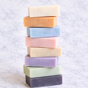 Op de afbeelding: Een stapel van acht rechthoekige zeepblokken in verschillende pastelkleuren, waaronder crème, oranje, lichtblauw, roze, beige, lavendel, groen en donkerpaars. De zepen zijn gerangschikt op een wit-grijs marmeren oppervlak.