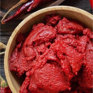 May include: A wooden bowl filled with vibrant red tomato paste, surrounded by dried red chili peppers. The peppers are arranged on a wooden surface, creating a rustic and appetizing display. The image showcases ingredients for cooking.