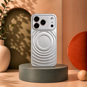Puede incluir: Una funda de smartphone plateada que presenta un recorte para cámara de triple lente con anillos dorados y un panel trasero texturizado con ondas circulares concéntricas. La funda tiene un acabado metálico y forma rectangular.