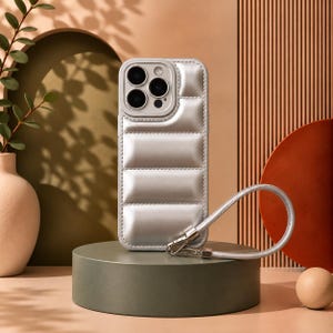 Puede incluir: Una funda de teléfono plateada, acolchada y estilo plumífero con textura guateada y una correa de muñeca a juego desmontable. La funda presenta un recorte elevado para la cámara y un acabado metálico suave y brillante.