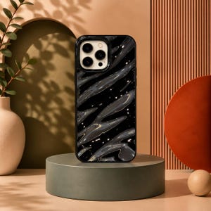 Puede incluir: Una funda de smartphone negra con un patrón de ondas plateadas brillantes y líquidas en la parte posterior. La funda tiene un recorte rectangular para la cámara en la esquina superior izquierda con tres aberturas circulares para lentes.