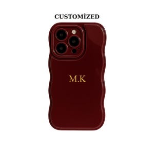 Może przedstawiać: Bordowa obudowa telefonu z falistym brzegiem. Obudowa ma wycięcie na aparat i inicjały "M.K" w złotych literach. Słowo "CUSTOMIZED" jest wydrukowane u góry. Obudowa ma chronić smartfon.