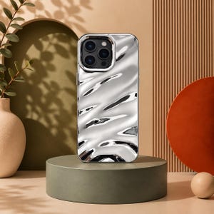 May include: A silver phone case with a reflective, liquid-like wavy surface texture. The case features a rectangular cutout for the camera module, which contains three circular lenses and a flash, all encased in a matching silver frame.