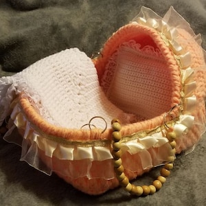May include: A crocheted baby bassinet with a white crocheted blanket and a white lace trim. The bassinet has a wooden handle and a peach color.