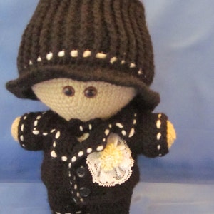 May include: A crocheted doll wearing a black hat and a black suit with white trim. The doll has a white lace collar and a yellow flower.