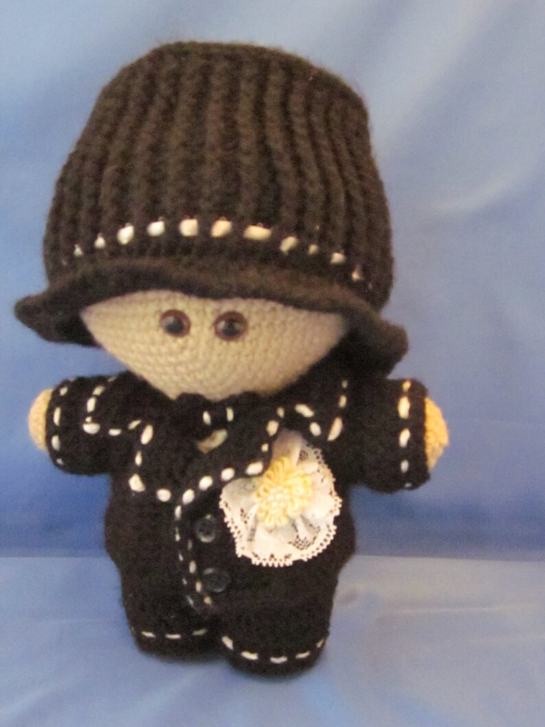 May include: A crocheted doll wearing a black hat and a black suit with white trim. The doll has a white lace collar and a yellow flower.