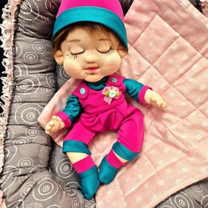 May include: A handmade doll dressed in a pink and teal outfit, including a hat and jumpsuit. The doll has closed eyes and is lying on a pink blanket with a star pattern, nestled in a gray patterned bed.