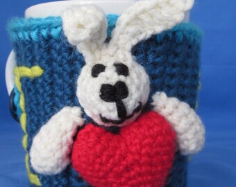 Op de afbeelding: Een gehaakte mokwarmer met een wit konijn dat een rood hart vasthoudt. De warmer is blauw met gele accenten.
