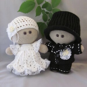 May include: Two crocheted dolls dressed in wedding attire. The bride doll wears a white crocheted dress with lace trim and a white crocheted hat with a flower. The groom doll wears a black crocheted suit with white buttons and a black crocheted hat.