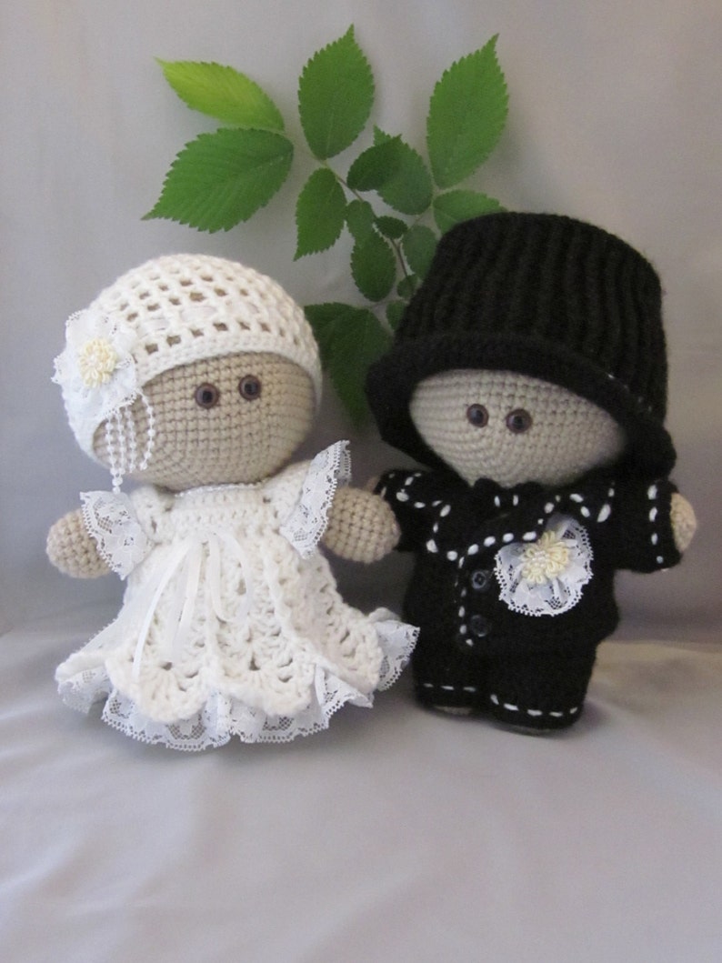 May include: Two crocheted dolls dressed in wedding attire. The bride doll wears a white crocheted dress with lace trim and a white crocheted hat with a flower. The groom doll wears a black crocheted suit with white buttons and a black crocheted hat.