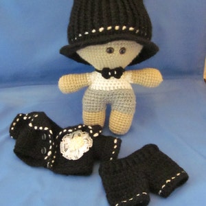May include: A crocheted doll wearing a black hat, a white shirt with a black bow tie, and a black jacket and pants. The doll has gray and white yarn for its body and black yarn for its hair.