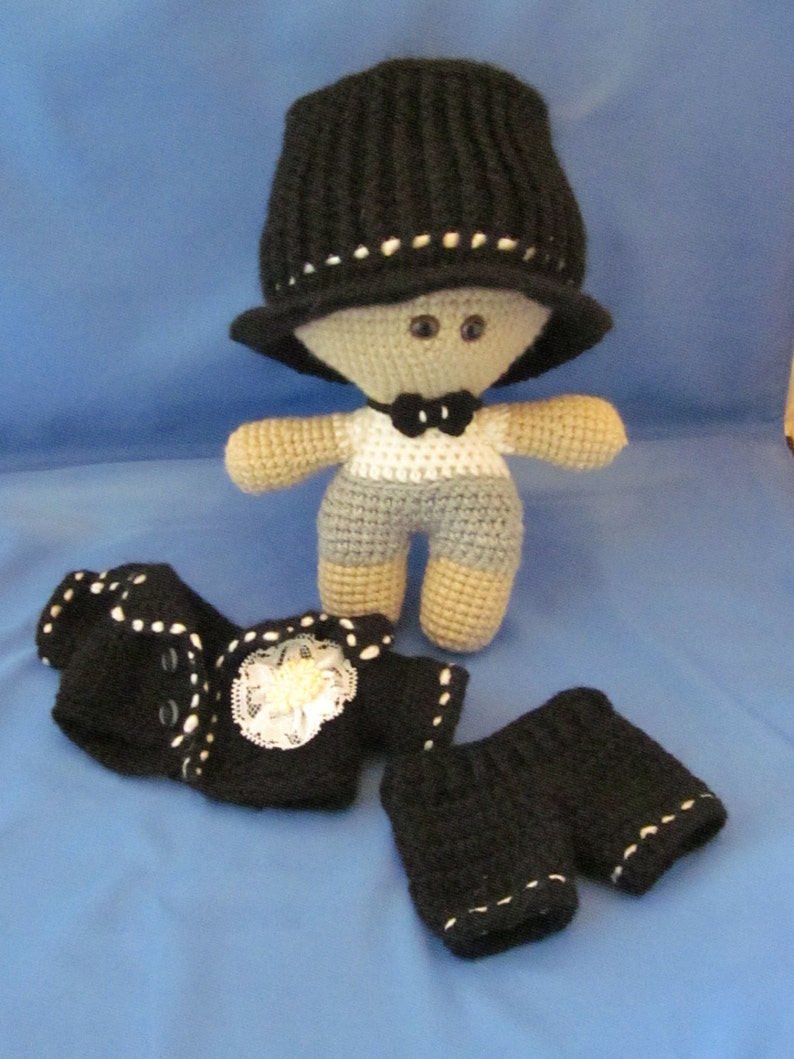 May include: A crocheted doll wearing a black hat, a white shirt with a black bow tie, and a black jacket and pants. The doll has gray and white yarn for its body and black yarn for its hair.