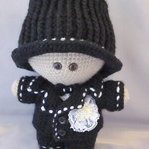 May include: A crocheted doll wearing a black top hat, a black suit with white trim, and a white lace bow. The doll has brown eyes and a beige face.