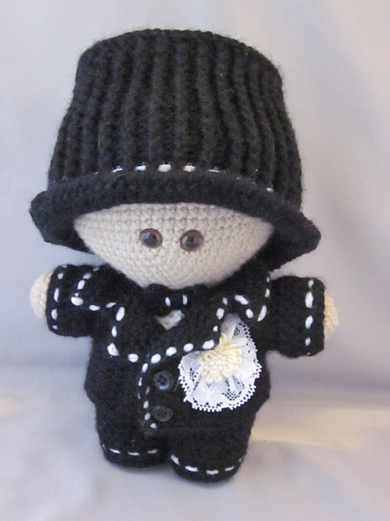 May include: A crocheted doll wearing a black top hat, a black suit with white trim, and a white lace bow. The doll has brown eyes and a beige face.