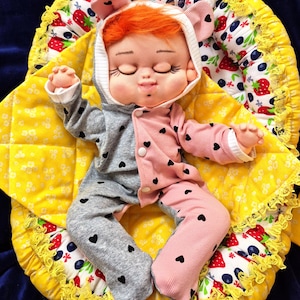 May include: A handmade doll with orange hair wearing a gray and pink outfit with black heart accents. The doll is lying on a yellow blanket with a strawberry and blueberry pattern, and a yellow lace trim. The doll is wearing a hooded cap with pink ears.