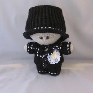 May include: A crocheted doll wearing a black top hat, a black jacket with white trim, and a white flower.