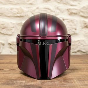 Pu&ograve; includere: Un casco Mandaloriano bordeaux con dettagli neri e le iniziali "M.F.C." sulla visiera. Il casco &egrave; realizzato in un materiale lucido e poggia su una superficie di legno. Il casco ha una caratteristica visiera a forma di T.