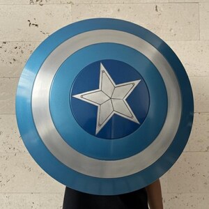 May include: A round shield with a blue, silver, and white color scheme. The shield features a central white star on a blue background, surrounded by silver and blue concentric circles. The shield is a prop or collectible.