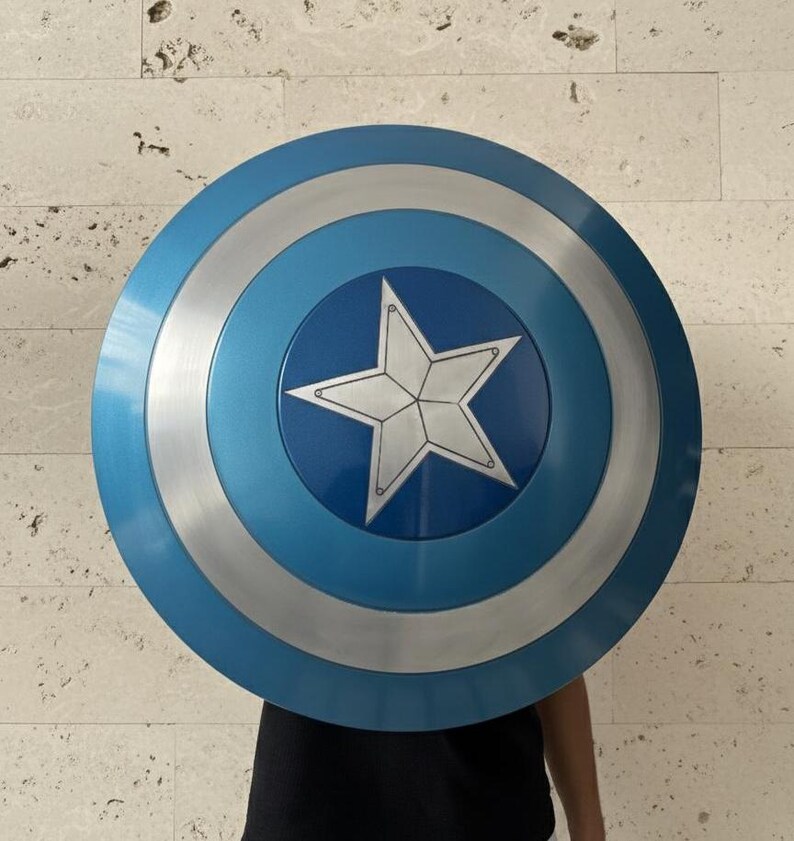 May include: A round shield with a blue, silver, and white color scheme. The shield features a central white star on a blue background, surrounded by silver and blue concentric circles. The shield is a prop or collectible.