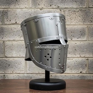 May include: A silver-colored medieval helmet with a visor and rivet detailing, displayed on a black stand. The helmet has a cylindrical shape with a flat top and a series of small holes on the visor. The text "medievalforgeco" is visible on the helmet.