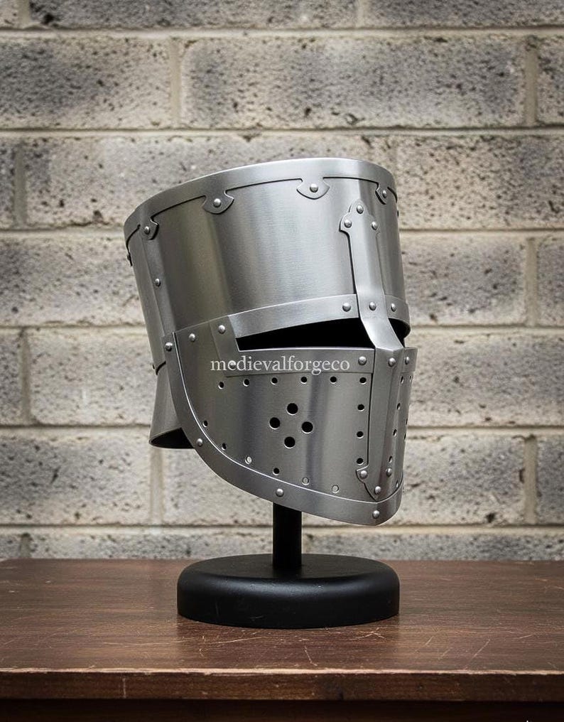 May include: A silver-colored medieval helmet with a visor and rivet detailing, displayed on a black stand. The helmet has a cylindrical shape with a flat top and a series of small holes on the visor. The text "medievalforgeco" is visible on the helmet.
