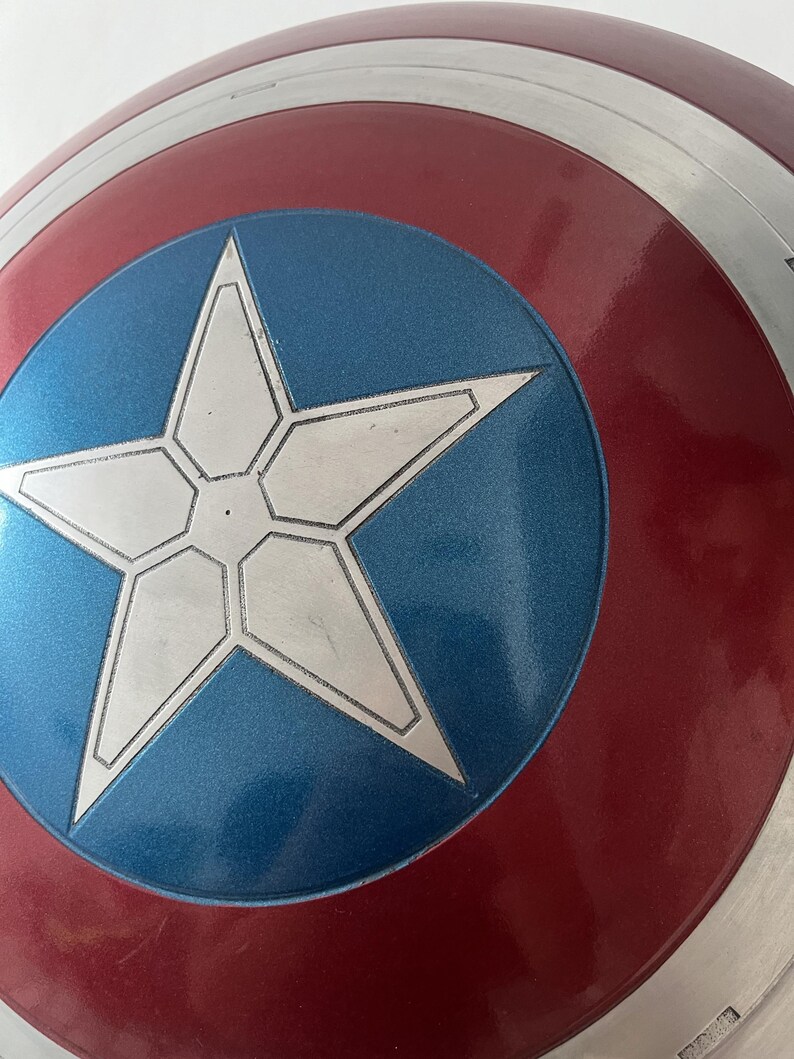 Puede incluir: Primer plano de un escudo del Capit&aacute;n Am&eacute;rica. El escudo es circular con un anillo exterior rojo y plateado. El centro presenta un c&iacute;rculo azul con una estrella blanca de cinco puntas. El escudo tiene un brillo met&aacute;lico.
