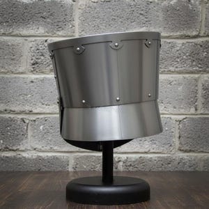 May include: A silver-colored medieval helmet, displayed on a black stand. The helmet has a rounded top and a flared lower section, with rivets along the seams. The background features a gray brick wall and a dark wooden surface.