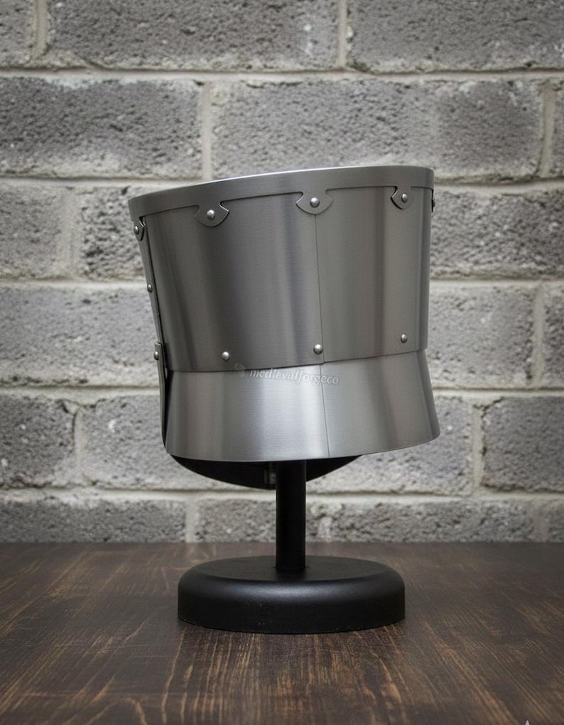 May include: A silver-colored medieval helmet, displayed on a black stand. The helmet has a rounded top and a flared lower section, with rivets along the seams. The background features a gray brick wall and a dark wooden surface.