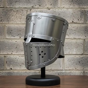 May include: A silver-colored medieval helmet with riveted construction, displayed on a black stand. The helmet features a visor with multiple small holes and is set against a brick wall background. The helmet is a historical replica.