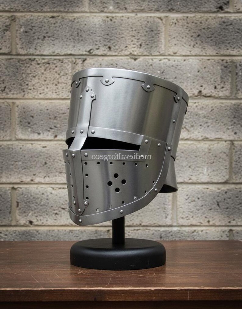 May include: A silver-colored medieval helmet with riveted construction, displayed on a black stand. The helmet features a visor with multiple small holes and is set against a brick wall background. The helmet is a historical replica.