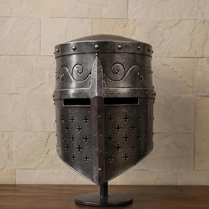 May include: A dark gray metal great helm featuring a flat top, a horizontal eye slit, and a lower section with a grid of small cross-shaped ventilation cutouts. The helmet is adorned with engraved scrollwork patterns and visible rivets.