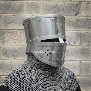 May include: A silver-colored medieval knight's helmet with a riveted design and a visor with small ventilation holes. The helmet is worn over a chainmail coif, offering protection for the head and neck. The background is a gray brick wall.
