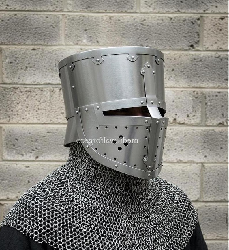 May include: A silver-colored medieval knight's helmet with a riveted design and a visor with small ventilation holes. The helmet is worn over a chainmail coif, offering protection for the head and neck. The background is a gray brick wall.