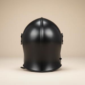 Pu&ograve; includere: Un elmo nero in stile medievale, con finitura liscia e opaca. L'elmo ha una parte superiore arrotondata e un bordo inferiore curvo, con piccoli dettagli metallici sui lati. Lo sfondo &egrave; beige neutro.
