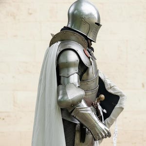 May include: A medieval knight's full suit of armor, including a helmet, chest plate, arm and leg protection, and gauntlets. The silver-colored armor is worn with a white cape and a small shield. A sword is visible at the knight's side.
