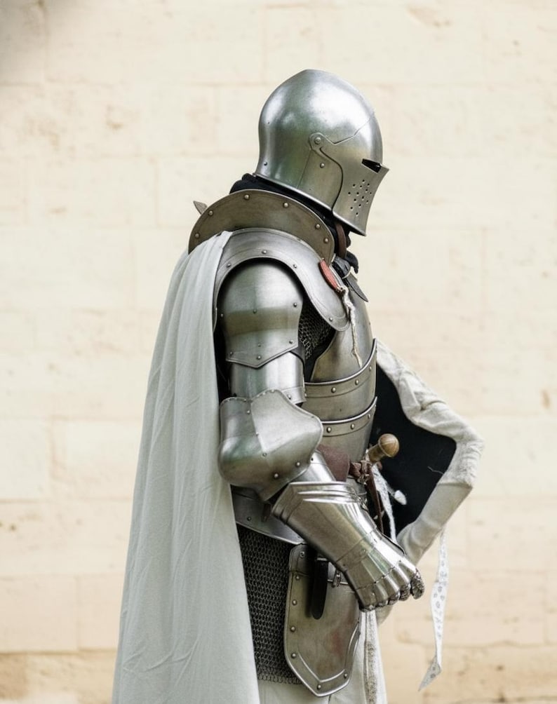 May include: A medieval knight's full suit of armor, including a helmet, chest plate, arm and leg protection, and gauntlets. The silver-colored armor is worn with a white cape and a small shield. A sword is visible at the knight's side.