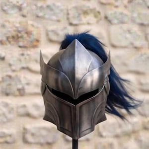 May include: A metallic silver knight helmet featuring a pointed visor, a horizontal eye slit, and a dark blue plume extending from the top. The helmet is mounted on a black vertical stand, showcasing a polished finish with visible contours.