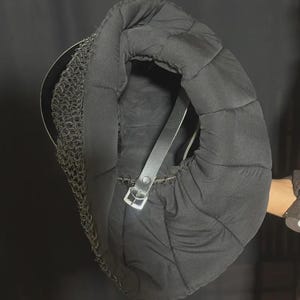 May include: A black fencing helmet with chainmail and a padded interior. The helmet has a black leather strap with a silver buckle. The chainmail covers the neck and lower face area. The padded interior is quilted.