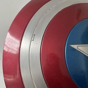 Puede incluir: Primer plano de un escudo del Capit&aacute;n Am&eacute;rica. El escudo es principalmente rojo con una banda plateada y una secci&oacute;n azul. El rojo tiene un brillo met&aacute;lico. Una estrella blanca es visible en la secci&oacute;n azul. El escudo tiene forma circular.
