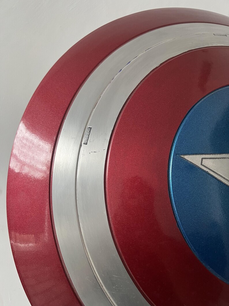 Puede incluir: Primer plano de un escudo del Capit&aacute;n Am&eacute;rica. El escudo es principalmente rojo con una banda plateada y una secci&oacute;n azul. El rojo tiene un brillo met&aacute;lico. Una estrella blanca es visible en la secci&oacute;n azul. El escudo tiene forma circular.