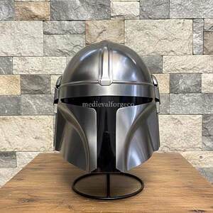 以下が含まれることがあります： T字型のバイザーが付いたメタリックシルバーのヘルメット。黒いスタンドに置かれています。ヘルメットは丸みを帯びた上部と角張った特徴があり、バイザーには「medievalforgeco」の文字が見えます。
