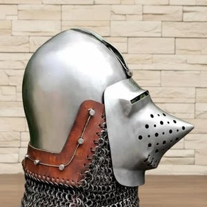 以下が含まれることがあります： 光沢のあるスチール製の騎士のヘルメット。尖ったバイザーと通気孔があります。茶色の革製のネックガードはリベットで固定され、鎖帷子で部分的に囲まれています。中世の鎧です。