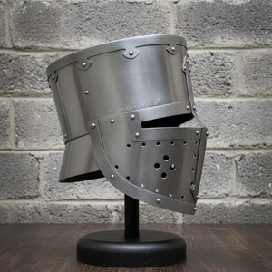 May include: A silver-colored medieval helmet with a visor, riveted construction, and a rounded top. The helmet is displayed on a black stand against a gray brick wall. The visor has several small holes.
