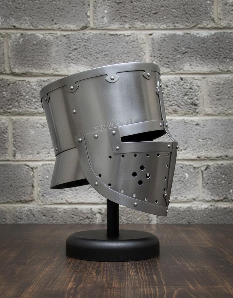 May include: A silver-colored medieval helmet with a visor, riveted construction, and a rounded top. The helmet is displayed on a black stand against a gray brick wall. The visor has several small holes.