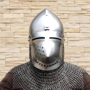 以下が含まれることがあります： 光沢のある銀色の、中世の兜。尖った上部と目のスリットがあります。ヘルメットには通気孔があり、鎖かたびらのフードが付いています。その人は茶色の衣服を着ています。