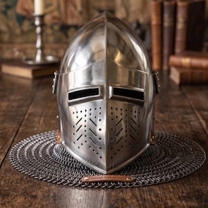 May include: A polished silver medieval helmet with a riveted visor and ventilation holes, resting on a chainmail coif. The helmet sits on a dark wooden table, with books and a candle holder in the background. A small tag reads "Authentic Medieval Armor."
